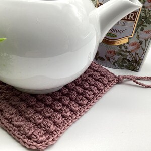 May include: A white ceramic teapot rests on a mauve crocheted square trivet. The trivet has a textured, raised pattern and a loop for hanging. A box of tea with floral designs is in the background.
