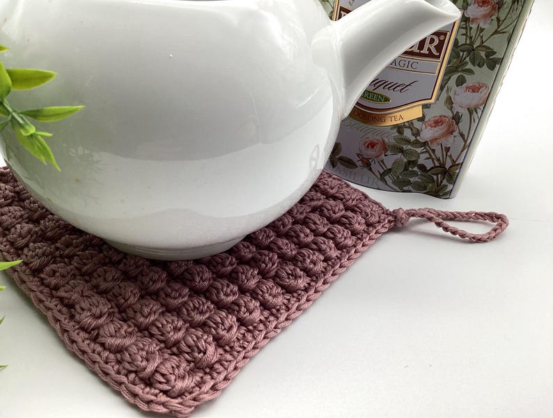 May include: A white ceramic teapot rests on a mauve crocheted square trivet. The trivet has a textured, raised pattern and a loop for hanging. A box of tea with floral designs is in the background.