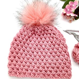 Könnte beinhalten: Eine rosafarbene Häkelmütze mit einem flauschigen rosa Bommel. Die Mütze ist mit einem strukturierten Stichmuster gearbeitet.