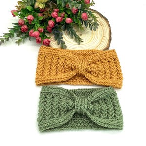 May include: Two knitted headbands in mustard yellow and sage green. The headbands feature a textured pattern and a knotted bow detail. They are displayed on a white surface, with a floral arrangement and a wooden slice in the background.