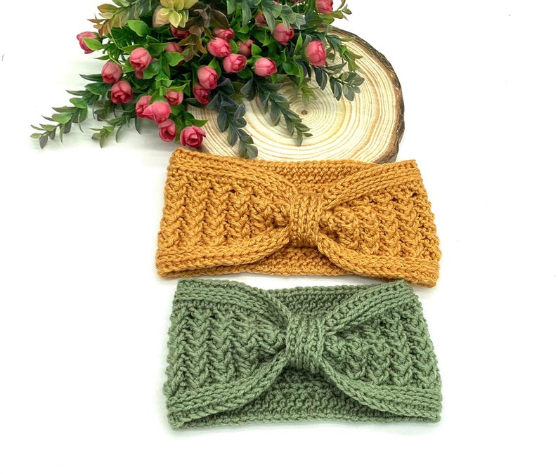May include: Two knitted headbands in mustard yellow and sage green. The headbands feature a textured pattern and a knotted bow detail. They are displayed on a white surface, with a floral arrangement and a wooden slice in the background.