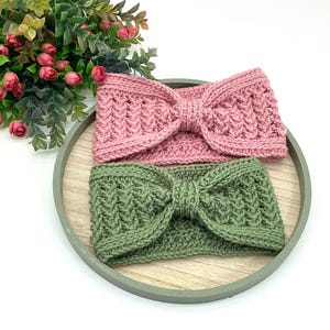 May include: Two knitted headbands are shown. One is a dusty pink, the other a muted green. Both have a textured pattern and a bow detail at the front. They are displayed on a wooden tray, with a plant in the background.