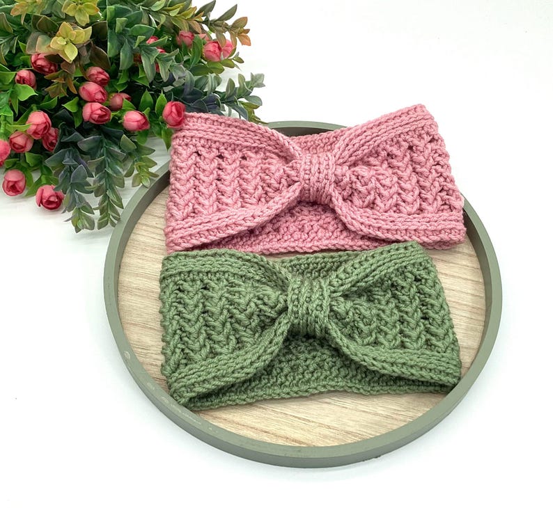May include: Two knitted headbands are shown. One is a dusty pink, the other a muted green. Both have a textured pattern and a bow detail at the front. They are displayed on a wooden tray, with a plant in the background.