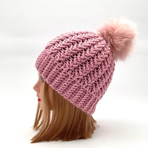 Może przedstawiać: R&oacute;żowa dzianinowa czapka typu beanie z teksturowanym wzorem i puszystym pomponem na g&oacute;rze. Czapka jest prezentowana na głowie manekina z blond włosami, podkreślając jej design i kolor.