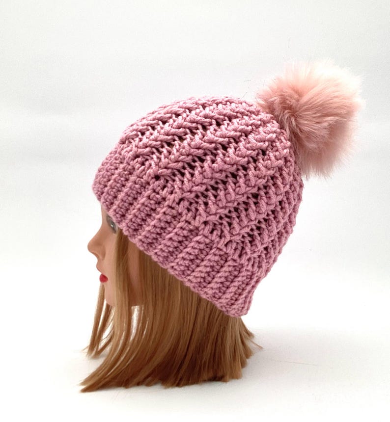 Może przedstawiać: R&oacute;żowa dzianinowa czapka typu beanie z teksturowanym wzorem i puszystym pomponem na g&oacute;rze. Czapka jest prezentowana na głowie manekina z blond włosami, podkreślając jej design i kolor.