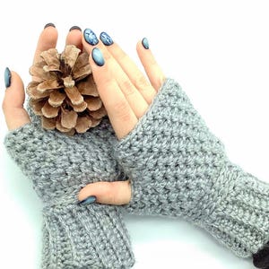 Könnte beinhalten: Ein Paar graue Fingerlose Handschuhe mit gerippter Textur. Die Handschuhe sind gehäkelt und haben eine Manschette, die sich bis zum Handgelenk erstreckt.