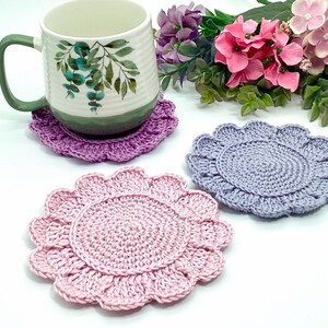 May include: A white ceramic mug with a green handle and botanical design sits on a purple crocheted coaster. Two additional crocheted coasters in pink and blue are also displayed. The coasters have a scalloped edge and a circular center.