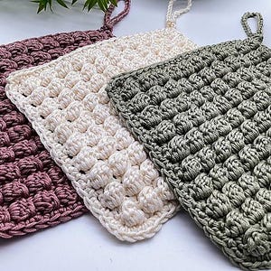 May include: Three handmade crochet potholders in shades of mauve, cream, and sage green. Each potholder features a textured, raised stitch pattern and a loop for hanging. These square potholders are perfect for kitchen use.
