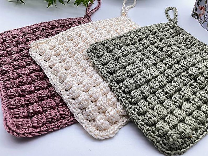 May include: Three handmade crochet potholders in shades of mauve, cream, and sage green. Each potholder features a textured, raised stitch pattern and a loop for hanging. These square potholders are perfect for kitchen use.