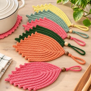 May include: A set of six leaf-shaped crochet pot holders in yellow, light green, pink, dark green, orange, and bright pink. Each piece features a matching crochet loop with a small wooden bead attached for hanging, arranged in a fan pattern.