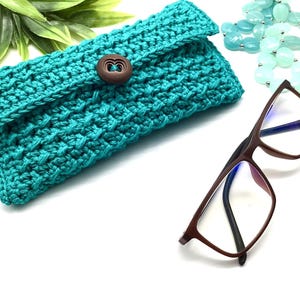 May include: A teal blue crocheted eyeglass case with a wooden button closure. The case is lying on a white surface next to a pair of brown eyeglasses.