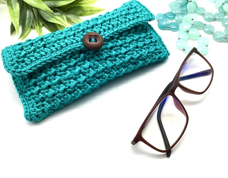 May include: A teal blue crocheted eyeglass case with a wooden button closure. The case is lying on a white surface next to a pair of brown eyeglasses.