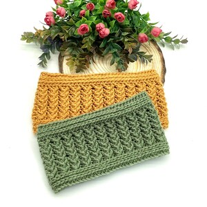 May include: Two knitted headbands in mustard yellow and sage green, with a textured cable knit pattern. The headbands are displayed on a white surface with a floral arrangement in the background, suggesting a handmade craft item.