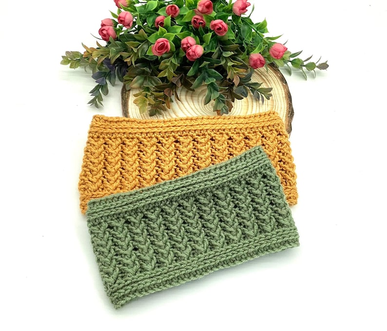May include: Two knitted headbands in mustard yellow and sage green, with a textured cable knit pattern. The headbands are displayed on a white surface with a floral arrangement in the background, suggesting a handmade craft item.