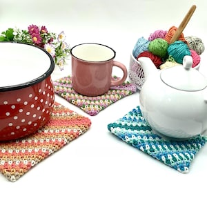 Puede incluir: Cuatro posavasos de ganchillo en diferentes colores, incluyendo rosa, verde, azul y naranja. Los posavasos están sobre una superficie blanca con una olla roja, una taza rosa y una tetera blanca.
