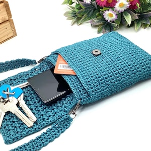 Pu&ograve; includere: Borsa a tracolla all'uncinetto color teal con chiusura a patta. La borsa &egrave; aperta, rivelando uno smartphone nero, chiavi e una carta. Anche la tracolla &egrave; color teal e all'uncinetto. La borsa &egrave; su una superficie bianca con fiori sullo sfondo.