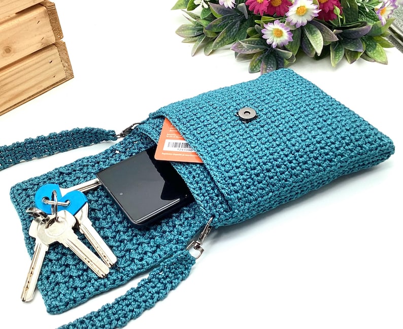 Pu&ograve; includere: Borsa a tracolla all'uncinetto color teal con chiusura a patta. La borsa &egrave; aperta, rivelando uno smartphone nero, chiavi e una carta. Anche la tracolla &egrave; color teal e all'uncinetto. La borsa &egrave; su una superficie bianca con fiori sullo sfondo.
