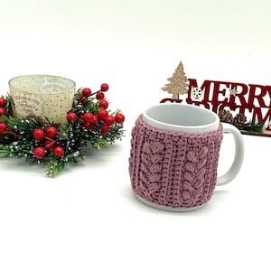 May include: A white ceramic mug with a mauve knitted cozy. The cozy has a cable knit design. A Christmas decoration with red berries and a candle holder is to the left. A wooden sign that says "Merry Christmas" is to the right.
