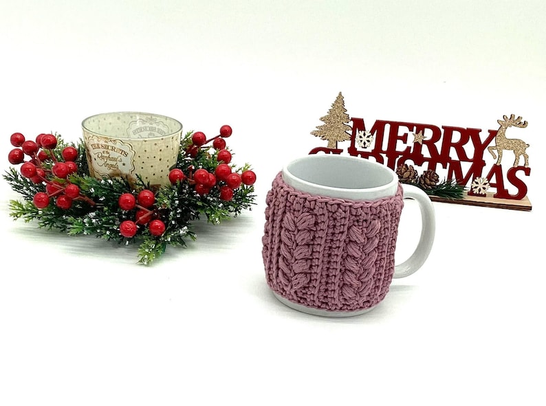 May include: A white ceramic mug with a mauve knitted cozy. The cozy has a cable knit design. A Christmas decoration with red berries and a candle holder is to the left. A wooden sign that says "Merry Christmas" is to the right.
