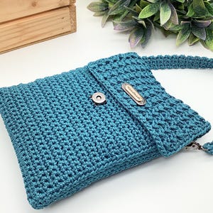 Pu&ograve; includere: Borsa a tracolla all'uncinetto color teal con chiusura a patta e chiusura magnetica argentata. La borsa presenta una tracolla abbinata e una piccola targhetta metallica con la scritta "Handmade".