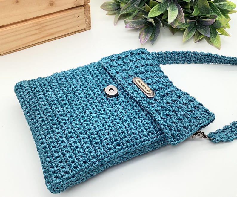 Pu&ograve; includere: Borsa a tracolla all'uncinetto color teal con chiusura a patta e chiusura magnetica argentata. La borsa presenta una tracolla abbinata e una piccola targhetta metallica con la scritta "Handmade".