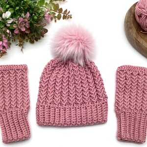 Op de afbeelding: Een bijpassende set roze gebreide winteraccessoires, waaronder een muts met een pluizige pompon en een paar vingerloze handschoenen. De muts en handschoenen hebben een textuur met een chevron-achtig patroon. De set wordt gepresenteerd op een wit oppervlak.