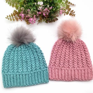 Puede incluir: Dos gorros de punto, uno turquesa y otro rosa, cada uno rematado con un pompón esponjoso. Los gorros tienen un patrón de chevron texturizado y un borde acanalado. Hojas verdes y flores en el fondo.