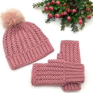 May include: A pink knitted hat with a faux fur pom-pom and matching fingerless gloves. The hat and gloves have a textured chevron pattern. The set is displayed on a white surface with a floral arrangement in the background.
