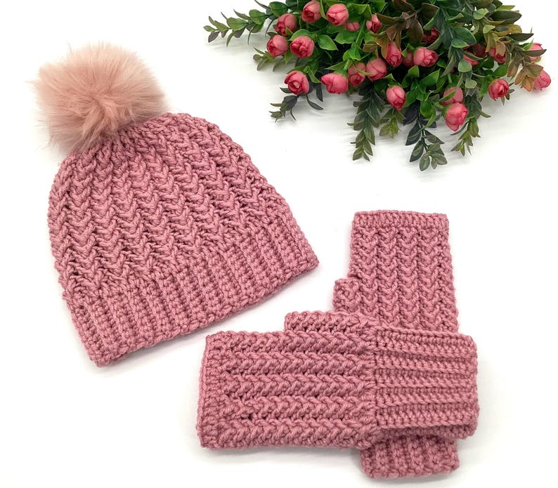 May include: A pink knitted hat with a faux fur pom-pom and matching fingerless gloves. The hat and gloves have a textured chevron pattern. The set is displayed on a white surface with a floral arrangement in the background.