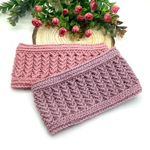 May include: Two hand-knitted headbands in shades of pink and mauve. The headbands feature a textured, cable-knit design. The background includes greenery and pink flowers, suggesting a handcrafted, seasonal theme.
