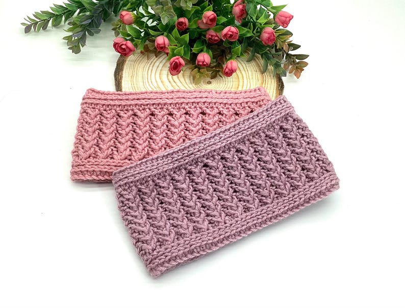 May include: Two hand-knitted headbands in shades of pink and mauve. The headbands feature a textured, cable-knit design. The background includes greenery and pink flowers, suggesting a handcrafted, seasonal theme.