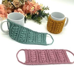 May include: A collection of handmade crochet mug cozies and a face mask in various colors. The mug cozies are mustard yellow and teal, while the face mask is pink. The crochet items feature a textured, raised stitch pattern and are designed to fit around mugs.