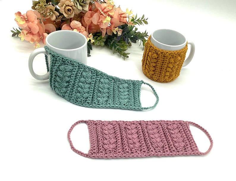 May include: A collection of handmade crochet mug cozies and a face mask in various colors. The mug cozies are mustard yellow and teal, while the face mask is pink. The crochet items feature a textured, raised stitch pattern and are designed to fit around mugs.