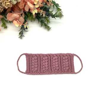 May include: A mauve-colored, crocheted face mask with a textured, vertical pattern. The mask has ear loops and is displayed on a white background with floral accents in the upper left corner. The mask is handmade.