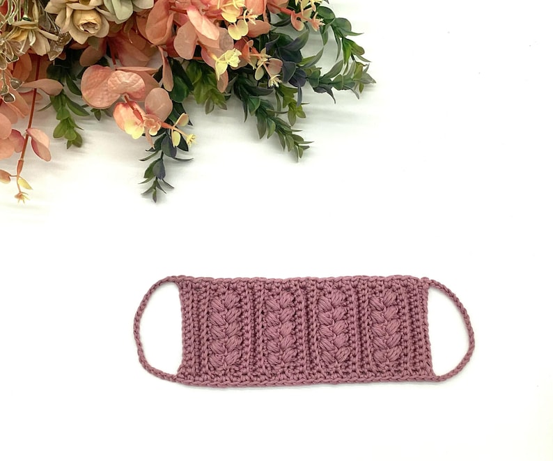 May include: A mauve-colored, crocheted face mask with a textured, vertical pattern. The mask has ear loops and is displayed on a white background with floral accents in the upper left corner. The mask is handmade.