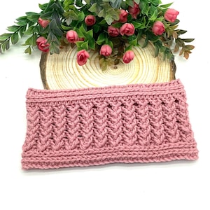 May include: A dusky pink knitted headband with a textured, chevron-like pattern. The headband is made of a thick yarn and has a ribbed top and bottom edge. The headband is displayed on a wooden slice with pink flowers.