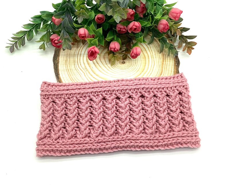 May include: A dusky pink knitted headband with a textured, chevron-like pattern. The headband is made of a thick yarn and has a ribbed top and bottom edge. The headband is displayed on a wooden slice with pink flowers.