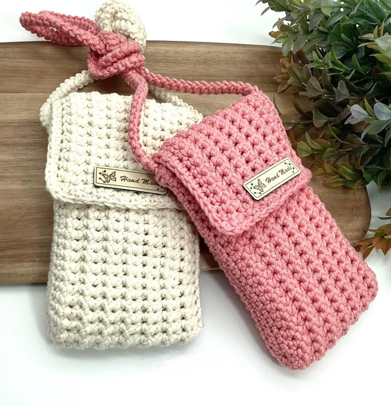 May include: Two crocheted phone pouches with adjustable straps. One pouch is cream coloured and the other is pink. Both pouches have a small wooden tag that says "Hand Made".