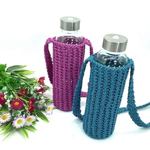 May include: Two glass water bottles with silver lids are encased in handmade crochet bottle holders. One holder is a deep purple, the other is teal. Both have matching straps. A bouquet of flowers sits to the left.