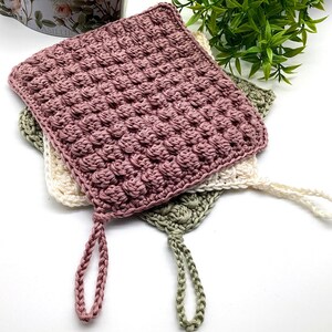 May include: Three handmade crochet washcloths in shades of mauve, cream, and sage green. Each square washcloth features a textured, raised pattern and a loop for hanging. The washcloths are stacked on top of each other.