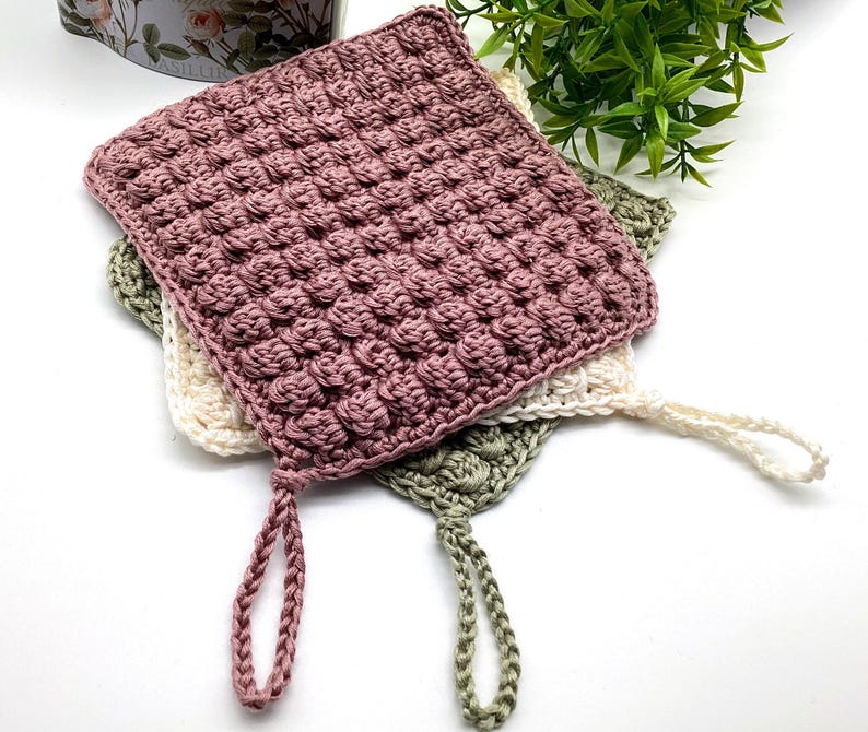 May include: Three handmade crochet washcloths in shades of mauve, cream, and sage green. Each square washcloth features a textured, raised pattern and a loop for hanging. The washcloths are stacked on top of each other.