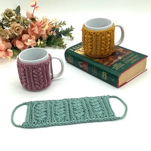 May include: Two white ceramic mugs with cable-knit cozies in mustard yellow and mauve. A matching seafoam green coaster is also present. The mugs are on a book and a white surface, with a bouquet of flowers in the background.