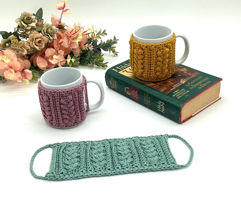 May include: Two white ceramic mugs with cable-knit cozies in mustard yellow and mauve. A matching seafoam green coaster is also present. The mugs are on a book and a white surface, with a bouquet of flowers in the background.