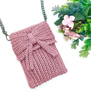 May include: A dusty rose crochet crossbody bag with a decorative bow closure. The bag features a silver chain strap and is displayed on a white background. The bag is approximately 7 inches tall and 5 inches wide.