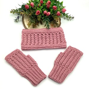 May include: A set of hand-knitted accessories in a dusty rose colour. The set includes a headband and two fingerless gloves. The items have a textured, woven pattern. A floral arrangement with pink roses sits in the background.