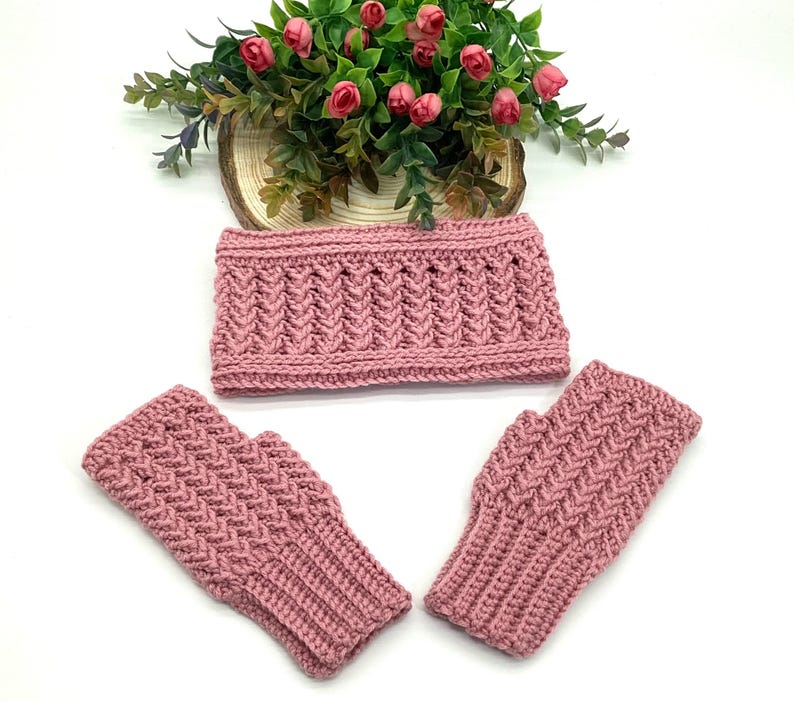 May include: A set of hand-knitted accessories in a dusty rose colour. The set includes a headband and two fingerless gloves. The items have a textured, woven pattern. A floral arrangement with pink roses sits in the background.