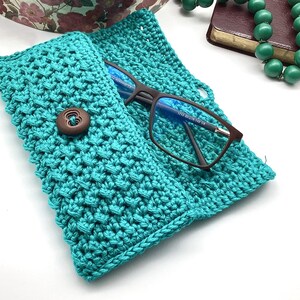 May include: A teal crocheted glasses case with a brown button closure. The case is open and a pair of eyeglasses is inside.