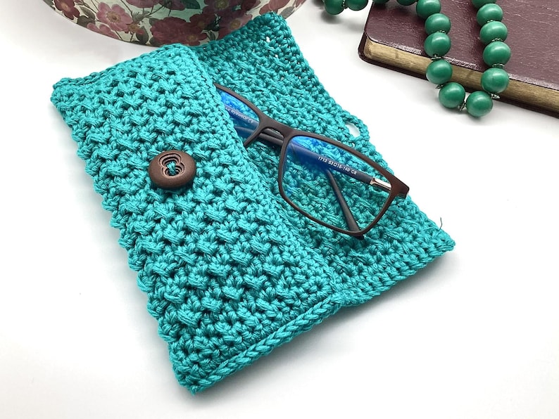 May include: A teal crocheted glasses case with a brown button closure. The case is open and a pair of eyeglasses is inside.