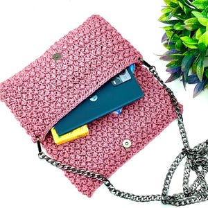 May include: A pink crocheted crossbody bag with a silver chain strap. The bag is open and a black smartphone is inside.