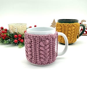 May include: Two mugs with crochet cozies. One mug has a pink cozy with a cable knit design, and the other has a yellow cozy. The mugs are on a white surface with holiday decorations in the background.